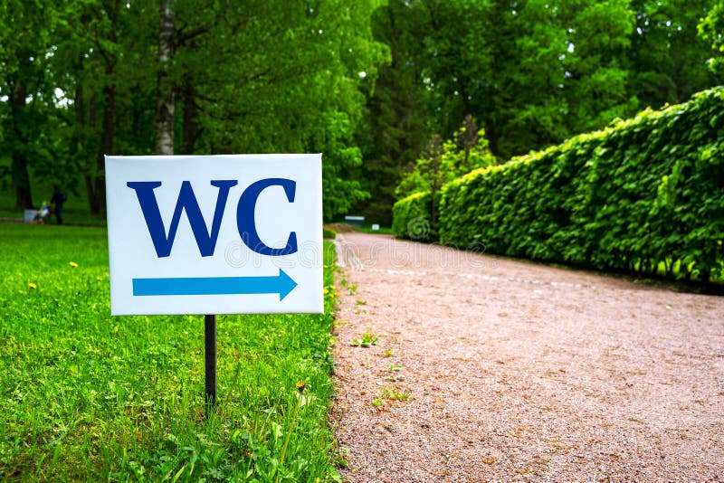 Toaletttecknet mot bakgrunden av gr?na tr?d i parkerar Vitt WC-tecken på plattan för vit metall med den indica blåa peka pilen arkivfoto Toaletttecknet mot bakgrunden av gr?na tr?d i parkerar Vitt WC-tecken på plattan för vit metall med den indica blåa peka pilen arkivfoto