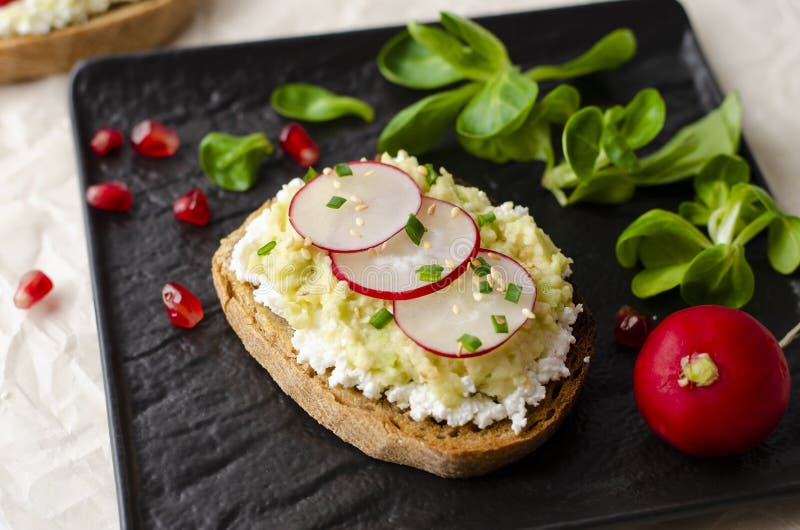 Toasts with cottage cheese and smashed avocado, radish, corn salad plant and pomegranate seeds on black plate. Top view. Copy