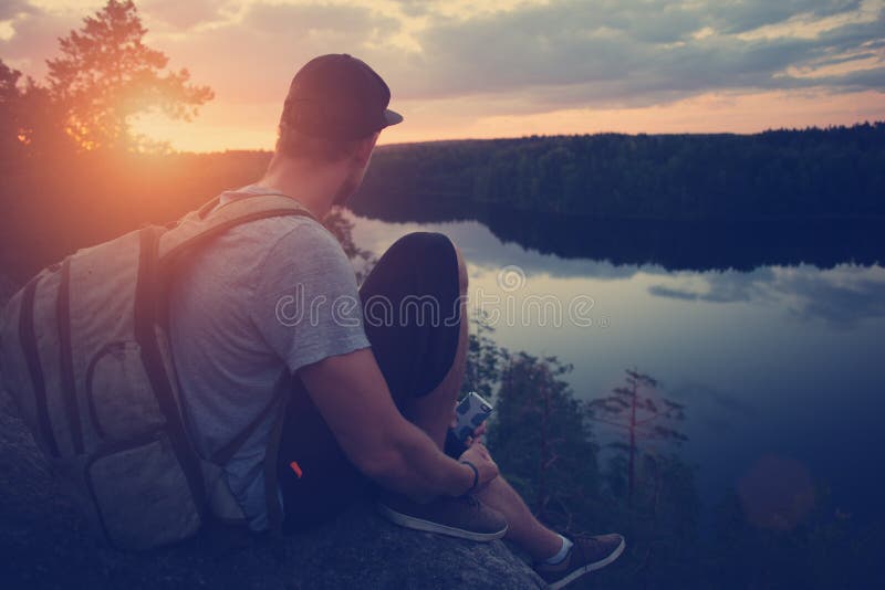 Young casual man with backpack sitting on the cliff edge above river and looking far away. Young casual man with backpack sitting on the cliff edge above river and looking far away