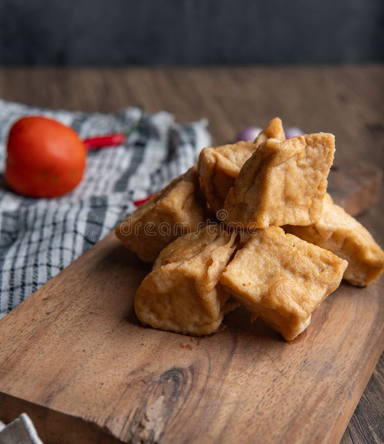 Tofu fresco frito en la tabla de corte de madera con especias y verduras. Tofu frito apilado en un tablero de madera rodeado de chile fresco, shallots y tomates en una mesa de cocina rústica. Perfecto para la cocina.
