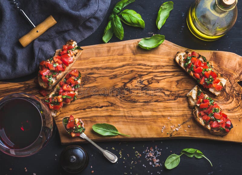 Tomato and basil bruschetta with glass of red wine, olive oil, salt, fresh herbs on wooden board over black background