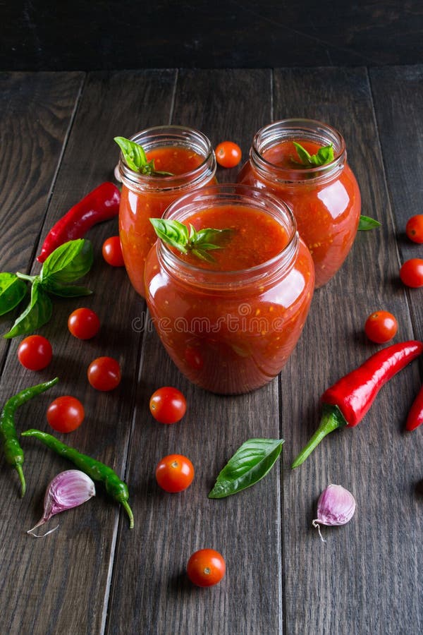 Tomato ketchup sauce with cherry tomatoes and red hot chili peppers, garlic and herbs in a glass jar on dark background.