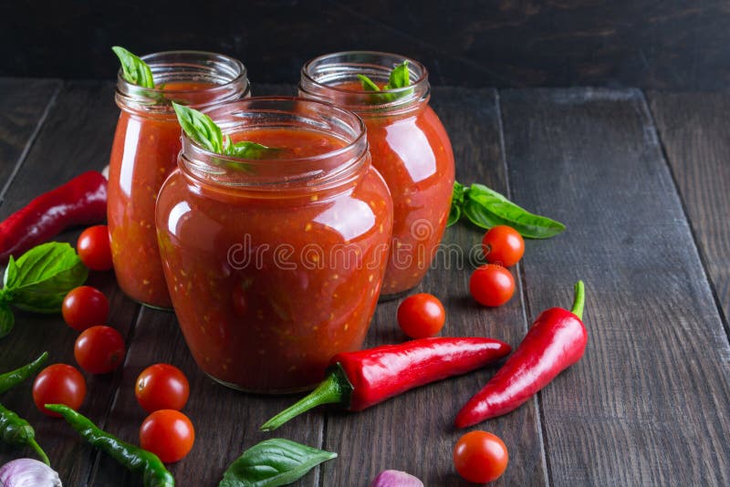 Tomato ketchup sauce with cherry tomatoes and red hot chili peppers, garlic and herbs in a glass jar on dark background.