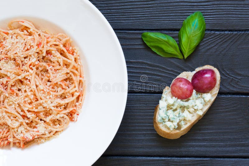 Tomato pasta with parmesan and bruschetta with pink grapes, basil and blue cheese on the black rustic wooden background. Italian