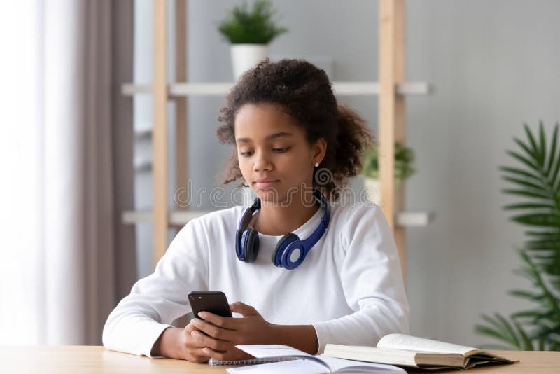 Focused African American teen girl using phone, distracted from homework, looking at screen, lazy schoolgirl chatting in social network instead of doing school tasks, children gadget addiction. Focused African American teen girl using phone, distracted from homework, looking at screen, lazy schoolgirl chatting in social network instead of doing school tasks, children gadget addiction