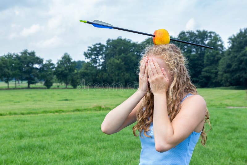 Teenage girl covering her face feeling fearful with apple and arrow on head in green meadow outdoors. Teenage girl covering her face feeling fearful with apple and arrow on head in green meadow outdoors