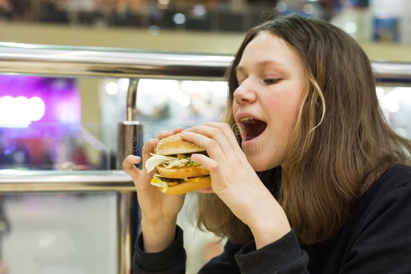 Tonårs- flicka som äter hamburgaren, mitt för bakgrundsshoppinggalleriaunderhållning royaltyfri foto Tonårs- flicka som äter hamburgaren, mitt för bakgrundsshoppinggalleriaunderhållning royaltyfri foto
