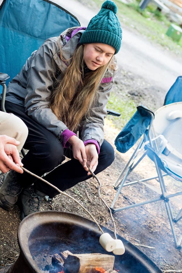 Teenage girl wearing beanie hat roasting large marshmallow on a stick over the campfire firepit. Camping family fun lifestyle. Teenage girl wearing beanie hat roasting large marshmallow on a stick over the campfire firepit. Camping family fun lifestyle