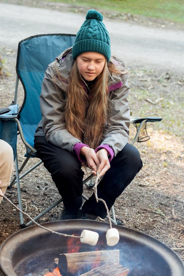 Teenage girl wearing beanie hat roasting large marshmallow on a stick over the campfire firepit. Camping family fun lifestyle. Teenage girl wearing beanie hat roasting large marshmallow on a stick over the campfire firepit. Camping family fun lifestyle