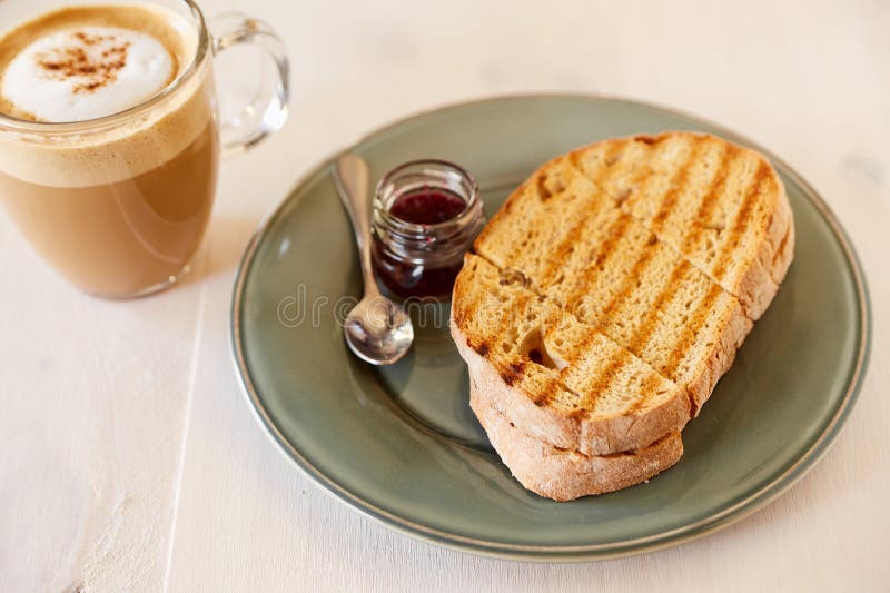 Toost met koffie en kersenjam op een witte houten lijst Gebraden brood op een grijze glanzende plaat Koffie met melk in een grote royalty-vrije stock afbeelding Toost met koffie en kersenjam op een witte houten lijst Gebraden brood op een grijze glanzende plaat Koffie met melk in een grote royalty-vrije stock afbeelding