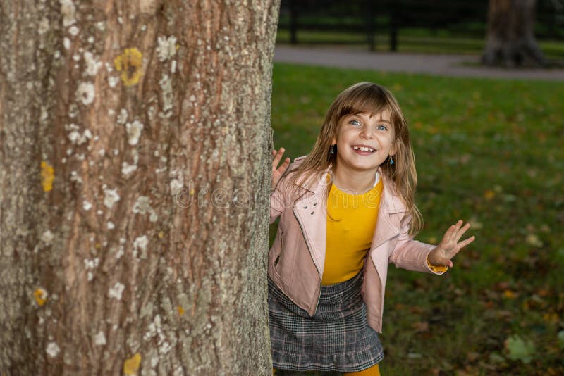 Photo of toothless funny cute kid girl playing hide and seek in the autumn Park. Looks out from behind a tree with a playful smile and laughs. Seasonal concept, happy childhood