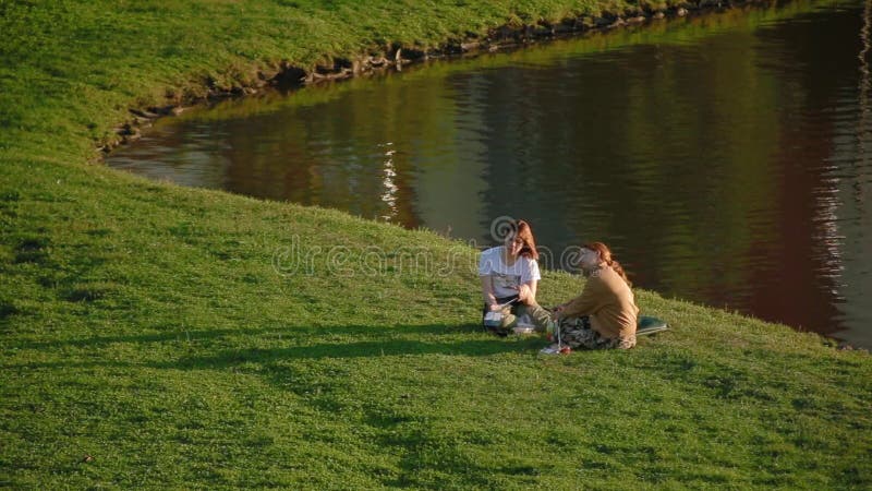 Two cute charming girls sit on green grass on lake and cute chats and eat