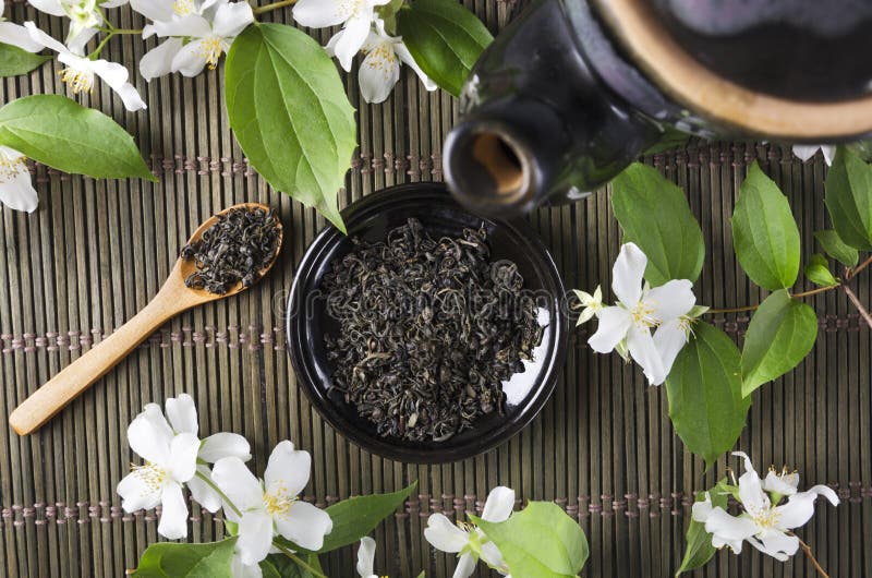 Top view of black teapot and saucer full of dry green tea, wooden spoon and aromatic fresh jasmine flowers on the green wicker mat stock photo Top view of black teapot and saucer full of dry green tea, wooden spoon and aromatic fresh jasmine flowers on the green wicker mat stock photo