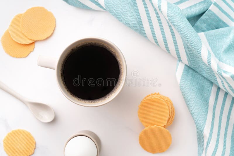 Top view of boiled egg in ceramic egg cup, cup of coffee and thin crispy corn chips on background of beautiful white