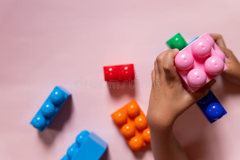 Top view on child&#x27;s hands playing with colorful plastic bricks on pink table. Kid building with geometric shapes stock photo