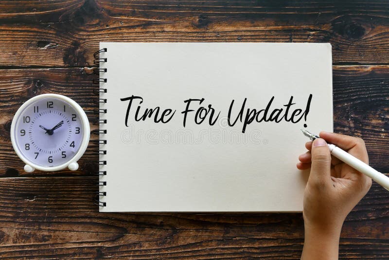 Top view of clock and hand holding pen writing Time For Update! on notebook on wooden background stock photo