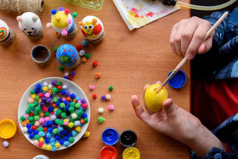 Top view of the desk and the hands of a child who paints festive easter eggs with  brushes