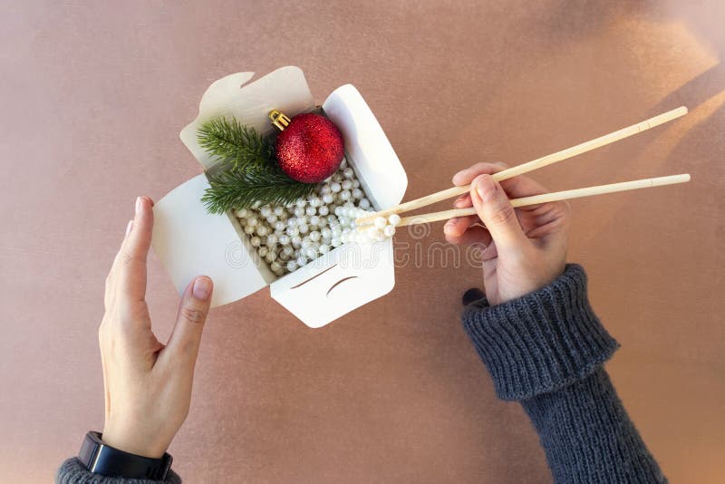 top view of female hands pull out white beads from a food box with Chinese chopsticks. red Christmas ball, branches and