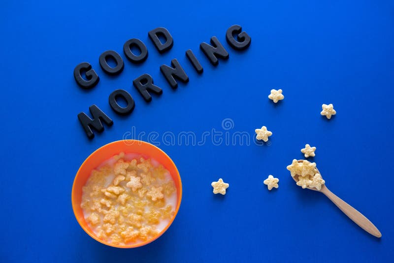 flat lay wooden bowl with crunchy corn stars and milk  and inscription good morning  on a blue background , space day