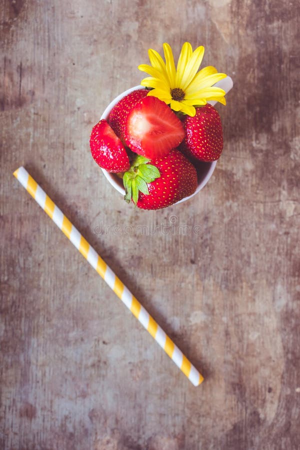 Top view of strawberries in a white cup and a yellow stripped straw on wooden background