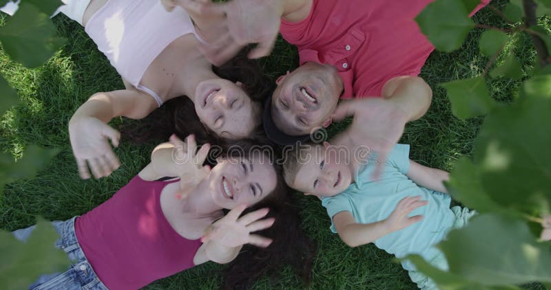 Top View of Happy Family Waving Hands at Camera and Smiling Laying Down in a Lawn under a Tree in the Summer