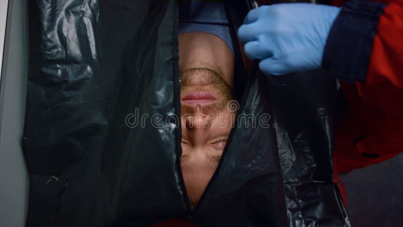 Top view medical worker hands closing black bag with dead men inside. Closeup paramedic in gloves zipping up body bag with corpse. Cadaver in plastic bag lying on stretchers in ambulance car. Top view medical worker hands closing black bag with dead men inside. Closeup paramedic in gloves zipping up body bag with corpse. Cadaver in plastic bag lying on stretchers in ambulance car