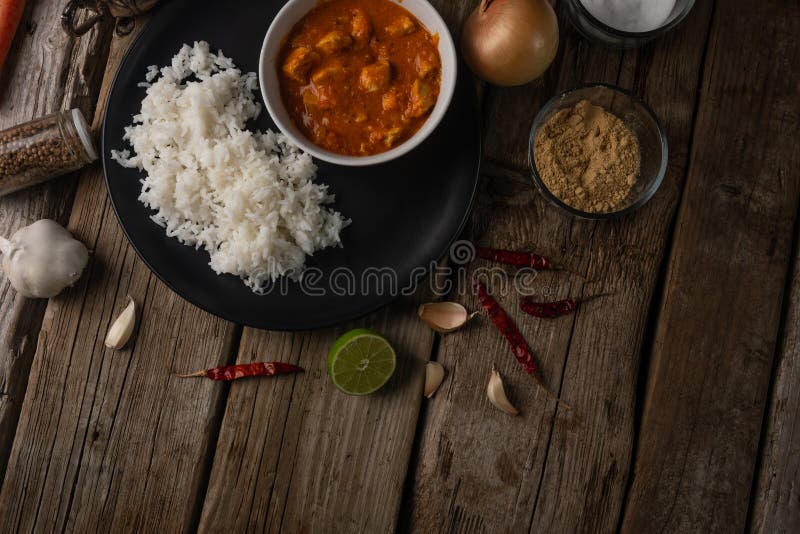 Top view of plate with classic chicken curry and rice served with lime, garlic and red hot chilli, onion and spices on rustic wooden table background. Traditional indian cuisine. Restaurant recipe.