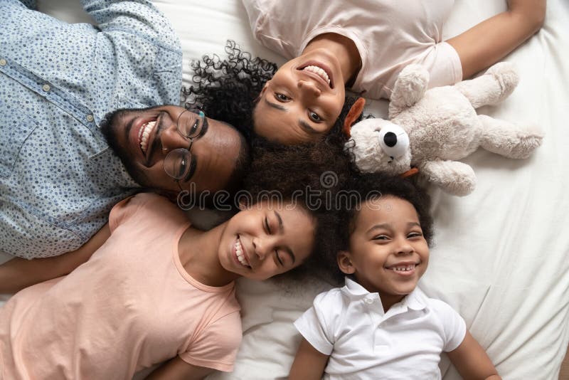 Top view portrait of happy young african American family with boy and girl kids lying on bed relaxing, smiling black parents rest with little daughter and son look at camera posing in home bedroom. Top view portrait of happy young african American family with boy and girl kids lying on bed relaxing, smiling black parents rest with little daughter and son look at camera posing in home bedroom