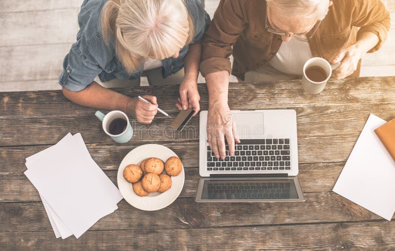 Top view of senior married couple paying for purchase in internet. They are sitting at desk and drinking tea with cookies. Top view of senior married couple paying for purchase in internet. They are sitting at desk and drinking tea with cookies