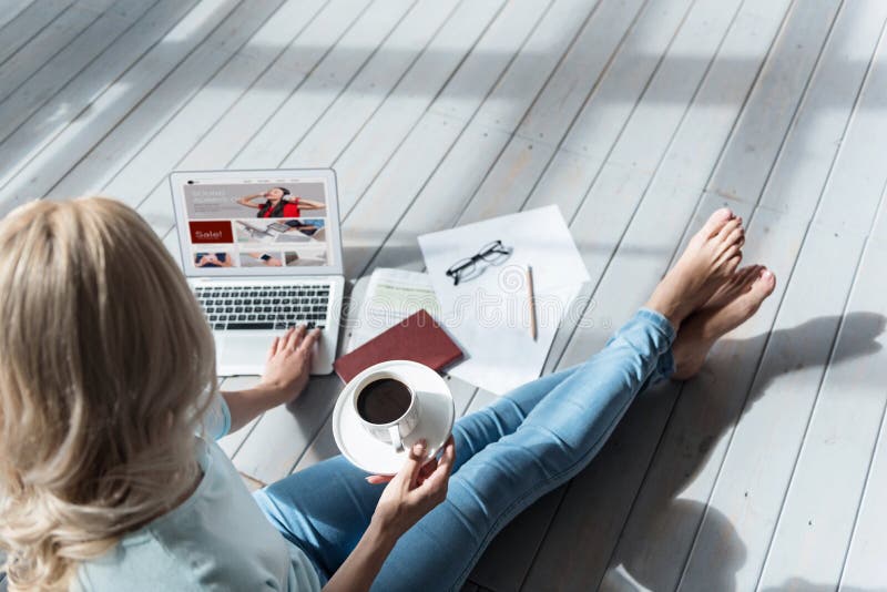 Weekend morning. Top view of blond woman sitting on grey wooden floor with full coffee cup and using her new laptop. Weekend morning. Top view of blond woman sitting on grey wooden floor with full coffee cup and using her new laptop.