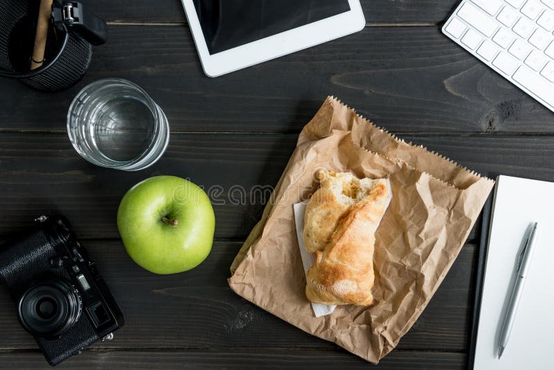 Top view of workplace with apple, bitten bun, glass of water and digital devices.