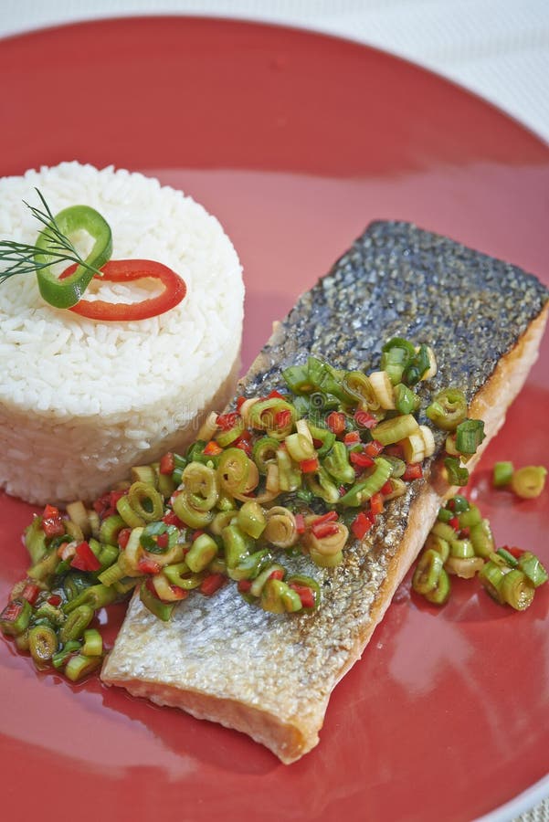 Torre de los pescados y del arroz en la placa roja. Pequeña torre del arroz llano al lado de un prendedero de pescados con la piel encendido cubierta en cebollas y chiles cortados en cuadritos Toda la sentada en una placa roja redonda grande