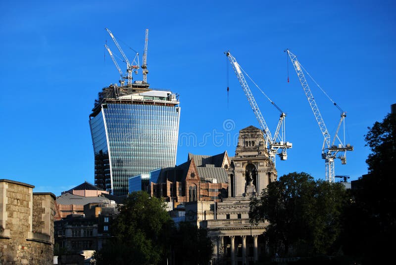 The Walkie Talkie tower at 20 Fenchurch Street under construction, seen with the old Port of London Authority in the foreground, blue sky, and tower cranes. The Walkie Talkie tower at 20 Fenchurch Street under construction, seen with the old Port of London Authority in the foreground, blue sky, and tower cranes.