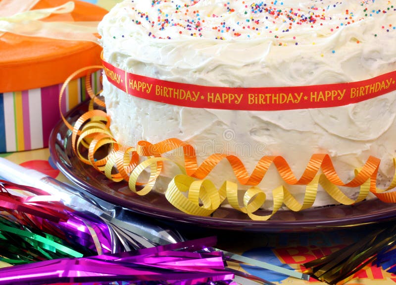 Vanilla birthday cake surrounded with orange and yellow ribbons and a Happy Birthday Ribbon all around. Party poppers and a gift by cake. Vanilla birthday cake surrounded with orange and yellow ribbons and a Happy Birthday Ribbon all around. Party poppers and a gift by cake.