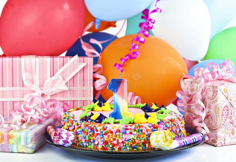 Pretty pink birthday cake with the number 1 candle. Cake is surrounded by gifts, party balloons, poppers and ribbons. Pretty pink birthday cake with the number 1 candle. Cake is surrounded by gifts, party balloons, poppers and ribbons.