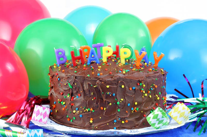 Chocolate cake decorated with Happy Birthday and surrounded with party poppers and streamers. Colorful balloons in the background with copy space. Chocolate cake decorated with Happy Birthday and surrounded with party poppers and streamers. Colorful balloons in the background with copy space.