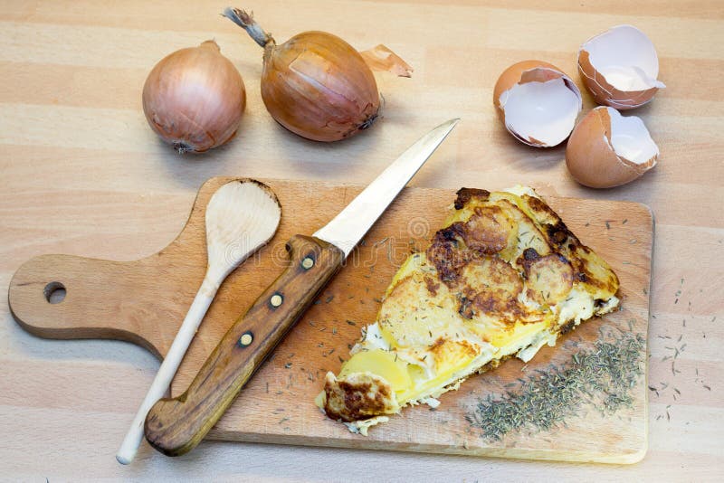 Slice of potato tortilla on wooden cutting board with egg shells, herbs and yellow onions. Slice of potato tortilla on wooden cutting board with egg shells, herbs and yellow onions.