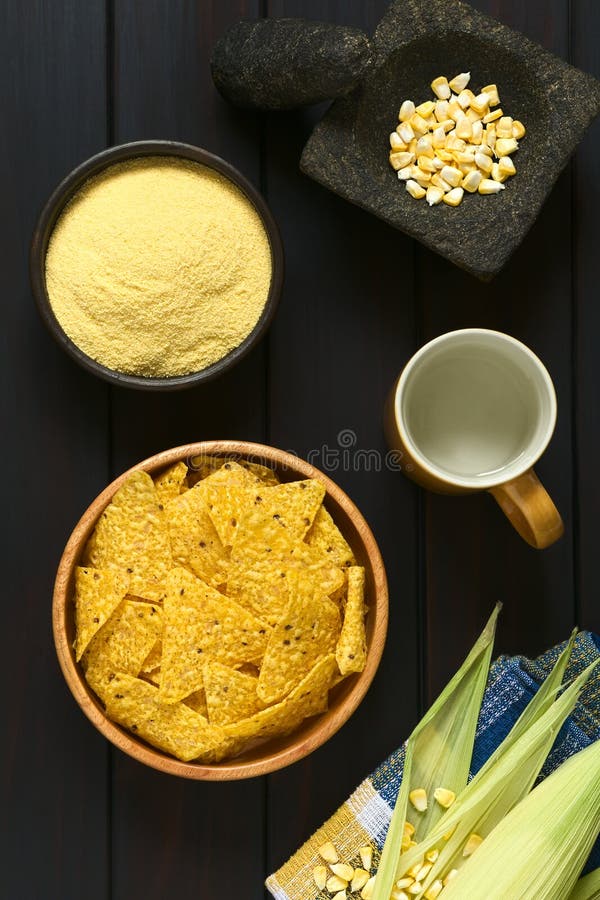 Overhead shot of tortilla chips in wooden bowl surrounded by its ingredients water, cornmeal and corn, photographed on dark wood with natural light. Overhead shot of tortilla chips in wooden bowl surrounded by its ingredients water, cornmeal and corn, photographed on dark wood with natural light