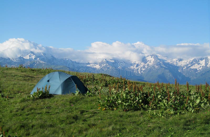 Tourist tent on the grass high in the mountains with beautiful rocky peaks covered with snow on the background