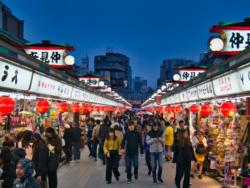 Tourists in the popular Nakamise Shopping Street near the Senso-ji Temple in Tokyo, Japan - taken at dusk showing the illuminated signs and lanterns of the tourist shops. Tourists in the popular Nakamise Shopping Street near the Senso-ji Temple in Tokyo, Japan - taken at dusk showing the illuminated signs and lanterns of the tourist shops.