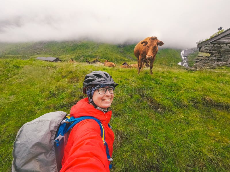 Touristischer Radfahrer der kaukasischen Frau im Sturzhelm und im Regenmantel nimmt selfie Foto gegen norwegische Berge und a lizenzfreie stockfotografie Touristischer Radfahrer der kaukasischen Frau im Sturzhelm und im Regenmantel nimmt selfie Foto gegen norwegische Berge und a lizenzfreie stockfotografie