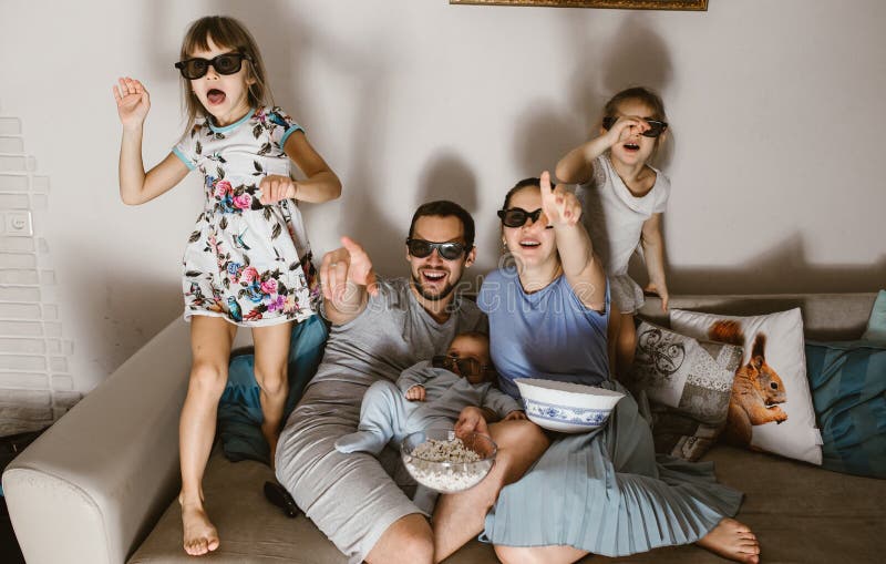 Tout le père de famille avec le bébé sur ses bras, mère et deux filles dans les verres spéciaux regardant la TV et mangeant du ma image stock Tout le père de famille avec le bébé sur ses bras, mère et deux filles dans les verres spéciaux regardant la TV et mangeant du ma image stock