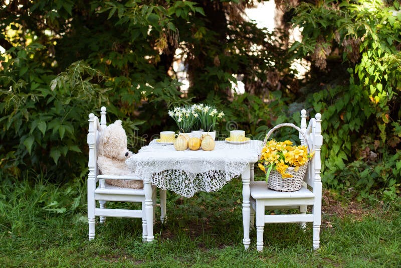 Toy Teddy bear sitting on white chair and cups of tea at the white small table. Children play area in garden. Outdoors wooden chai