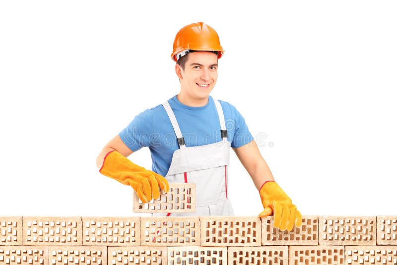 Trabajador de construcción de sexo masculino que sostiene un ladrillo detrás de la pared de ladrillo foto de archivo libre de regalías Trabajador de construcción de sexo masculino que sostiene un ladrillo detrás de la pared de ladrillo foto de archivo libre de regalías