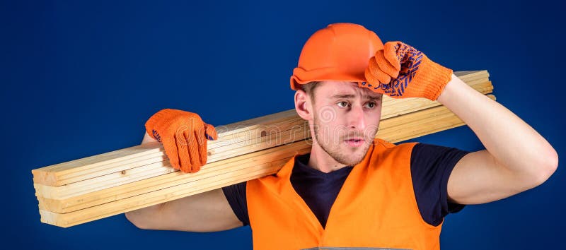 Trabajador de construcción que lleva un tablón de madera en su hombro Casco que lleva del carpintero, guantes protectores y segur foto de archivo Trabajador de construcción que lleva un tablón de madera en su hombro Casco que lleva del carpintero, guantes protectores y segur foto de archivo
