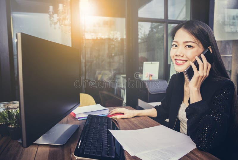 Asian working woman using computer in home office and talking on mobile phone with happiness face. Asian working woman using computer in home office and talking on mobile phone with happiness face