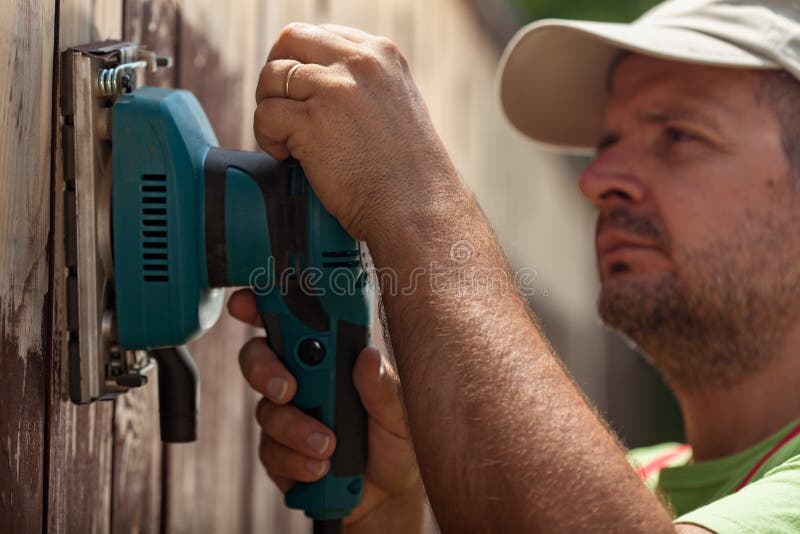 Trabalhador que usa uma máquina de lixar de vibração em uma cerca de madeira imagens de stock Trabalhador que usa uma máquina de lixar de vibração em uma cerca de madeira imagens de stock
