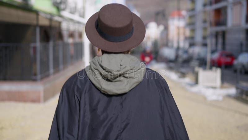 Tracking shot of confident young slim woman walking on city street. Back view of Caucasian lady in hat strolling