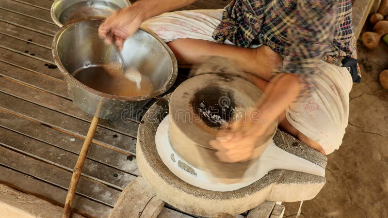 Woman sat cross-legged using hand-turned millstone to grind wet rice for making soaked rice flour