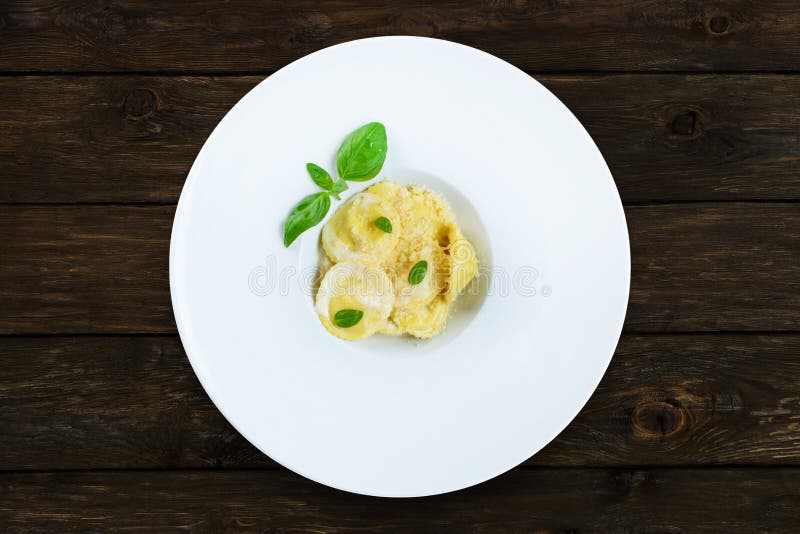 Traditional italian cuisine: ravioli with cream sause and parmesan decorated with basil on white round plate closeup - diet and healthy food at wooden table, above view. Traditional italian cuisine: ravioli with cream sause and parmesan decorated with basil on white round plate closeup - diet and healthy food at wooden table, above view